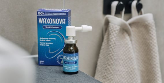 En bild av en flaska "Waxnova Wax Removal" med spraymunstycke och dess förpackning, placerad på en mörk yta. Förpackningen är blå och visar en öronillustration. I bakgrunden hänger en ljus handduk på en krok.