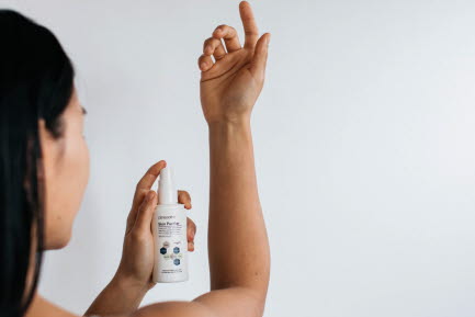 En person med långt, mörkt hår sprayar en produkt från en vit flaska på sin arm mot en ljus bakgrund.