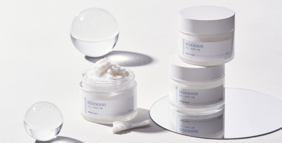 Två burkar med Mixsoon hudkräm staplade ovanpå varandra och en öppen burk med kräm på en spegelplatta. Runt produkterna finns dekorativa genomskinliga sfärer.