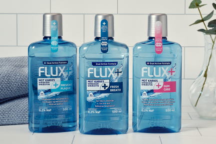 Tre Flux+ munsköljprodukter (Prevent Plaque, Fresh Breath och Gum Care) på en vit kakelbakgrund tillsammans med en handduk och en vas med växt.