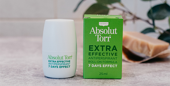 Både innerförpackning och ytterförpackning från Absolut Torr roll -on placerade bredvid varandra mot en grå bänkskiva med struktur. I bakgrunden syns en grön kvist och en fast tvål.