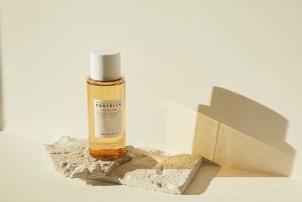 En flaska med Skin1004 Centella hudvårdsprodukt står på ett stenblock, framför en ljus beige bakgrund. Flaskan har en enkel etikett med vit text.