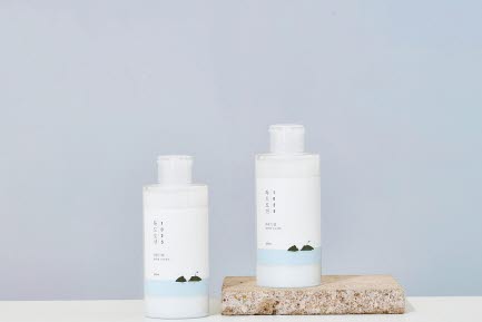 Två RoundLab hudvårdsflaskor står på ett korkblock framför en mjuk gradientbakgrund i blått och ljusrosa. Flaskorna har minimalistiska etiketter med bergsikoner och en ljusblå färgdetalj.