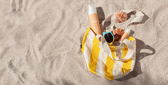 En gul och vitrandig strandväska fylld med ett par produkter från Beachkind på stranden med en snäcka i bakgrunden.