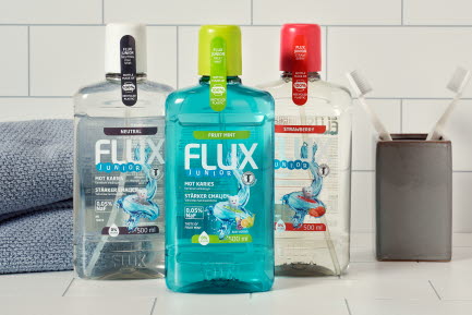 Tre Flux Junior munsköljprodukter (Neutral, Fruit Mint och Strawberry) på en vit kakelbakgrund tillsammans med en handduk och en tandborstkruka.